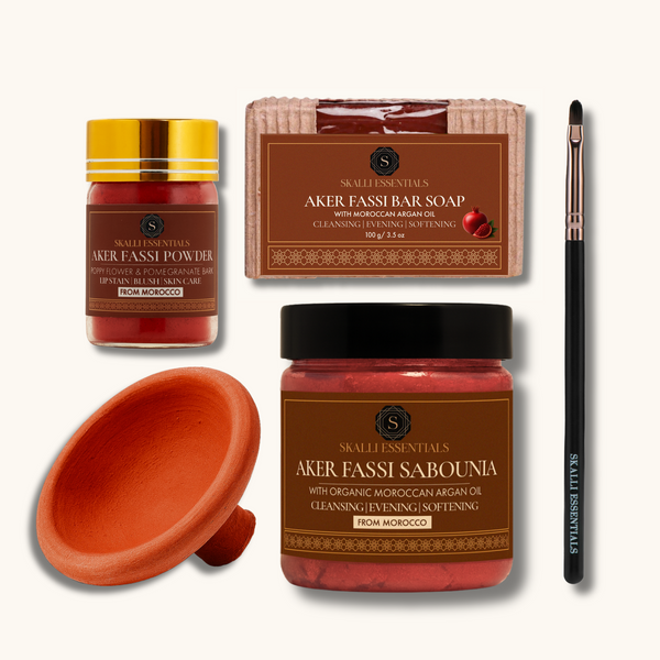 A Moroccan beauty set from Skalli Essentials on an off-white background, featuring a jar of Aker Fassi Sabounia, a jar of Aker Fassi Powder, a bar of soap, a terracotta aker fassi clay pot, and a black makeup brush.